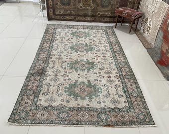 May include: Rectangular area rug with a cream-colored field and a central medallion design in shades of green, pink, and brown. The rug features a detailed border and fringe along the edges, showcasing an intricate pattern. The rug is placed on a tiled floor.