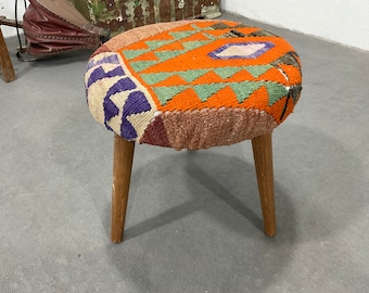 May include: A small, round, wooden stool with three legs. The seat is covered in a colorful, patterned fabric with orange, green, purple, and brown hues.