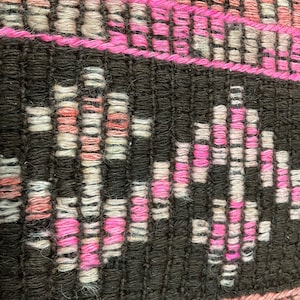 May include: Close-up of a woven textile with a geometric pattern. The design features alternating bands of black, pink, and off-white threads. The pink threads form a zig-zag pattern within the black bands, creating a textured surface.
