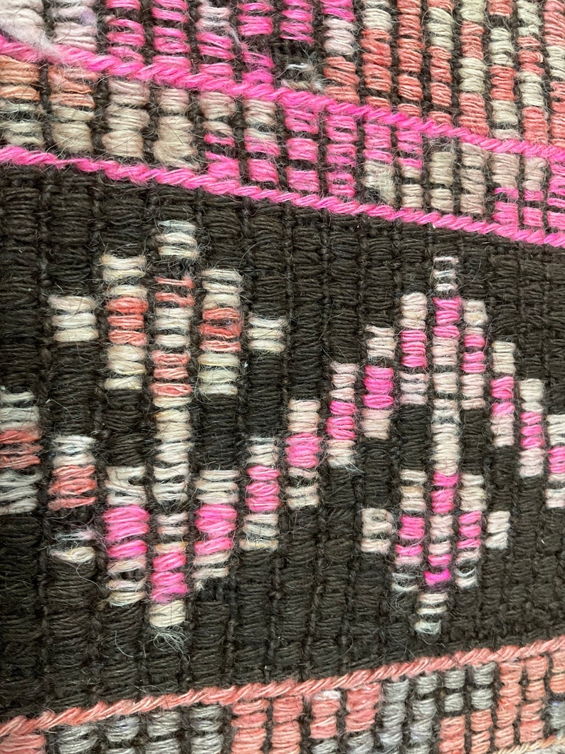 May include: Close-up of a woven textile with a geometric pattern. The design features alternating bands of black, pink, and off-white threads. The pink threads form a zig-zag pattern within the black bands, creating a textured surface.