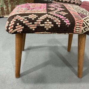 May include: A small, handcrafted stool with a patterned textile seat in shades of brown, pink, and white. The seat rests on four light-colored wooden legs. The stool appears to be a decorative or functional piece of furniture.