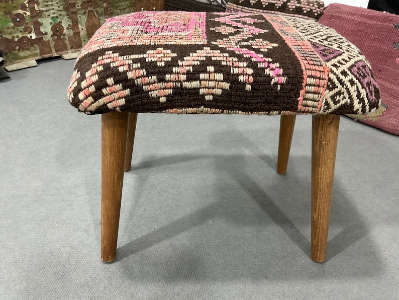 May include: A small, handcrafted stool with a patterned textile seat in shades of brown, pink, and white. The seat rests on four light-colored wooden legs. The stool appears to be a decorative or functional piece of furniture.