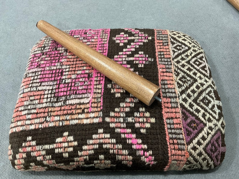 May include: A rectangular, upholstered ottoman with a brown, pink, and white patterned design. A wooden furniture leg is placed on top. The ottoman's fabric features geometric and floral motifs, creating a textured appearance.