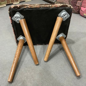 May include: A square ottoman with a patterned top in shades of pink, brown, and black. The ottoman has four wooden legs, each attached with metal brackets. The legs are angled outwards, providing stability. The underside of the ottoman is black.