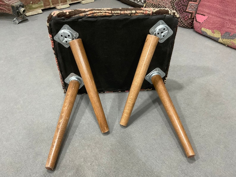 May include: A square ottoman with a patterned top in shades of pink, brown, and black. The ottoman has four wooden legs, each attached with metal brackets. The legs are angled outwards, providing stability. The underside of the ottoman is black.