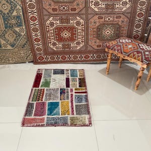 May include: A colorful patchwork rug with a variety of colors and patterns, including red, blue, green, and yellow. The rug is laid out on a white tiled floor. A wooden stool with a patterned cushion sits in the background.
