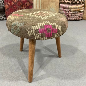 May include: A brown and pink patterned ottoman with three wooden legs. The ottoman is made of woven fabric and has a round top.