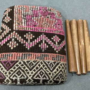 May include: A woven ottoman with a geometric pattern in shades of brown, pink, and white. The ottoman is accompanied by four wooden legs, ready for assembly. The design features a mix of textures and colors.