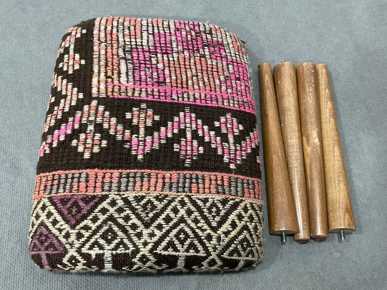 May include: A woven ottoman with a geometric pattern in shades of brown, pink, and white. The ottoman is accompanied by four wooden legs, ready for assembly. The design features a mix of textures and colors.