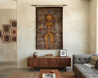 Puede incluir: Un tapiz rectangular que presenta una figura humana estilizada de color amarillo dorado con cabeza circular y extremidades alargadas, sobre un fondo marrón oscuro texturizado, suspendido de una barra metálica horizontal en la pared.