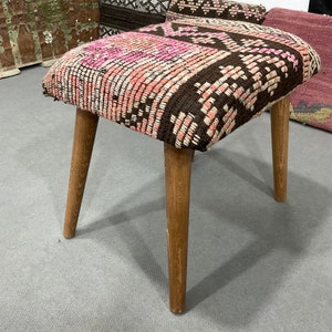 May include: A small, square stool with a patterned seat in shades of pink, brown, and white. The seat rests on four angled wooden legs. The stool is a decorative piece of furniture.