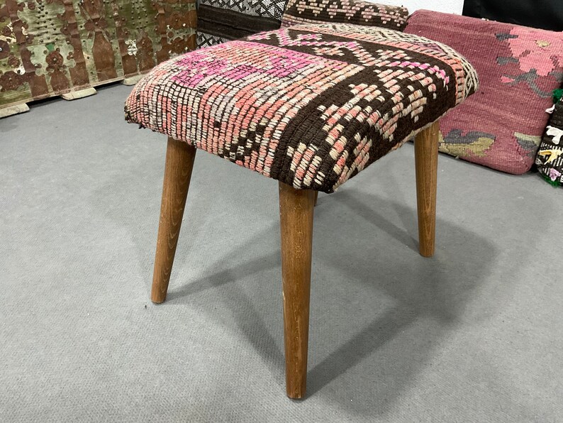 May include: A small, square stool with a patterned seat in shades of pink, brown, and white. The seat rests on four angled wooden legs. The stool is a decorative piece of furniture.