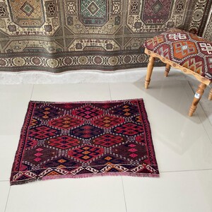 May include: A small, rectangular rug with a geometric pattern in shades of red, purple, and orange. The rug is placed on a white tiled floor. A small wooden stool with a patterned top is in the background.