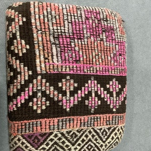 May include: A rectangular woven textile with a repeating pattern of pink, white, and brown threads. The edges are bordered with a darker brown and a geometric design in white and purple.