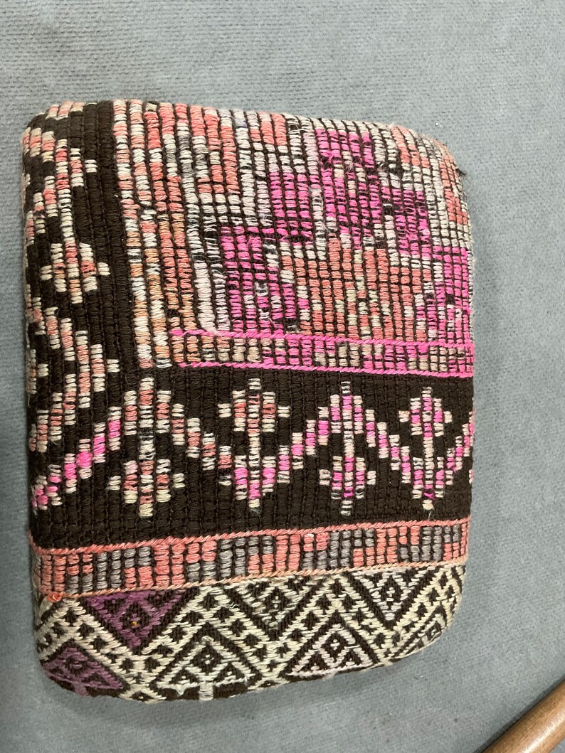 May include: A rectangular woven textile with a repeating pattern of pink, white, and brown threads. The edges are bordered with a darker brown and a geometric design in white and purple.