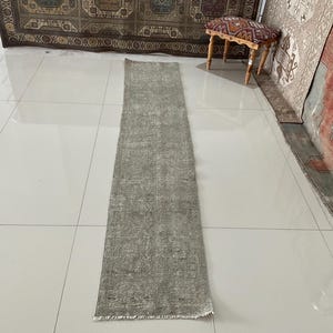 May include: A rectangular, light green runner rug with a faded, distressed appearance. The rug has a central design with a border and fringe on the short ends. The rug is placed on a tiled floor.