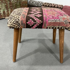 May include: A small, upholstered stool with a rectangular seat and four wooden legs. The seat features a patterned design with brown, pink, and cream colors. The legs are a light brown color.