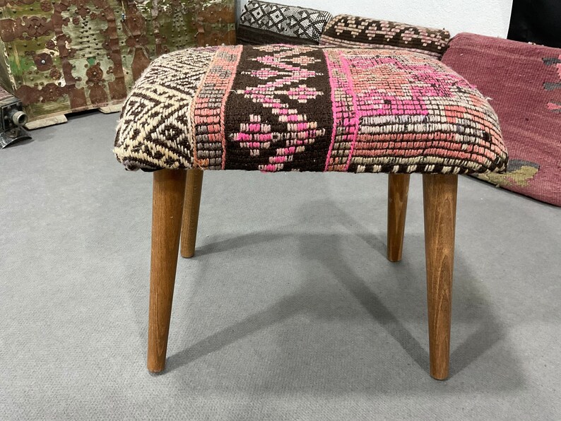 May include: A small, upholstered stool with a rectangular seat and four wooden legs. The seat features a patterned design with brown, pink, and cream colors. The legs are a light brown color.