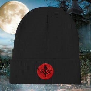 Op de afbeelding: Zwarte beanie met een rood cirkelvormig embleem met een gestileerd boomontwerp. De hoed is gemaakt van gebreid materiaal en heeft een omgeslagen rand. Het embleem is op de voorkant geborduurd.