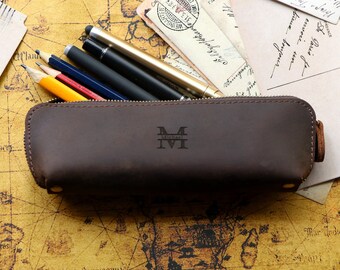 May include: A brown leather pencil case with a zipper closure. The case has the initials "M.M." embossed on the front. The case is filled with pencils and pens.