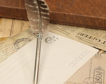 Puede incluir: Un bolígrafo plateado con una pluma de pluma unida a la parte superior. El bolígrafo está sobre una postal vintage con una caja de cuero marrón en el fondo.