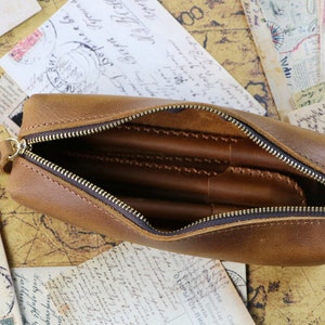 May include: A brown leather pencil case with a zip closure. The case is open and has several pens inside.