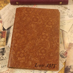 May include: A tan leather notebook cover featuring an embossed floral pattern across the entire front surface. The bottom right corner of the cover displays the text Love JMT in a dark, clear, sans-serif font. The cover has a rectangular shape.