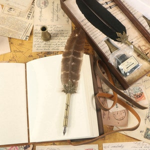 Puede incluir: Un diario encuadernado en cuero marr&oacute;n con una p&aacute;gina en blanco abierta, una pluma de pluma marr&oacute;n con una punta dorada y un dise&ntilde;o de hoja de metal, y una correa de cuero marr&oacute;n. El diario est&aacute; sobre una mesa con un mapa y otros papeles.