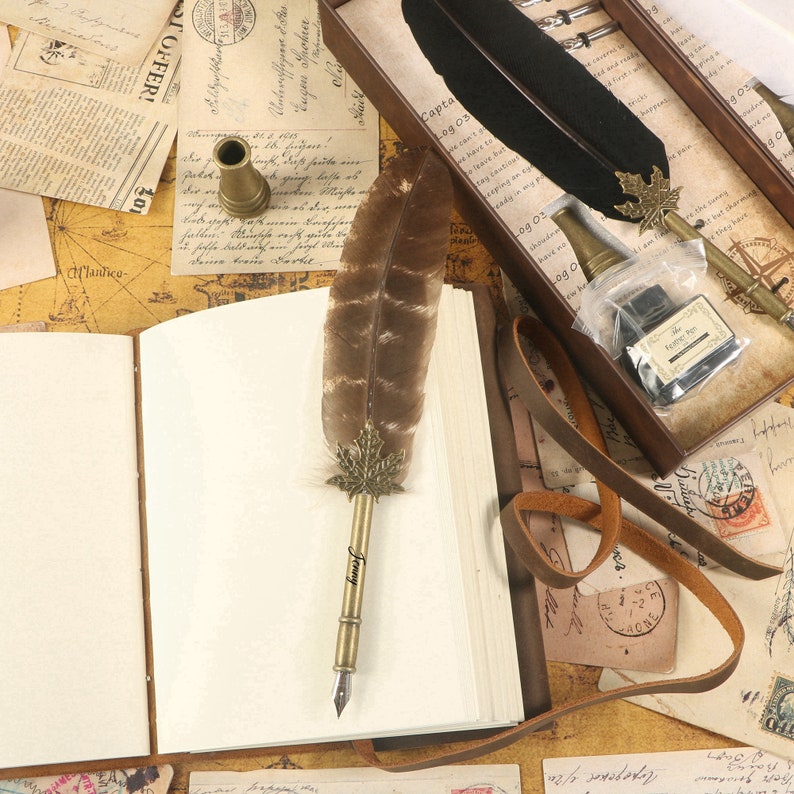 Puede incluir: Un diario encuadernado en cuero marr&oacute;n con una p&aacute;gina en blanco abierta, una pluma de pluma marr&oacute;n con una punta dorada y un dise&ntilde;o de hoja de metal, y una correa de cuero marr&oacute;n. El diario est&aacute; sobre una mesa con un mapa y otros papeles.