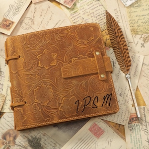 May include: A brown leather journal with an embossed floral design. The journal has a strap closure and the initials "IPSM" are written on the cover. A feather quill pen rests beside the journal on a background of vintage postcards.