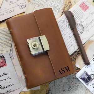 Puede incluir: Un diario de cuero marrón con un cierre de latón y una cerradura. El diario tiene las iniciales "ASM" grabadas en la portada.