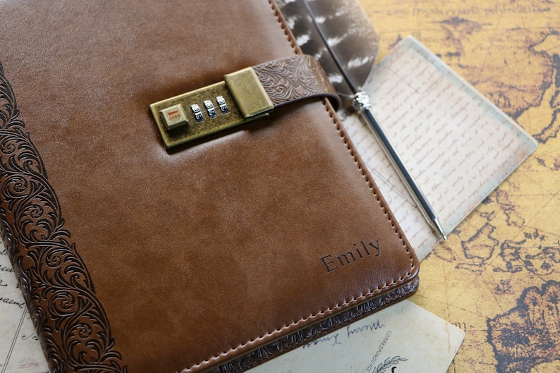 Op de afbeelding: Een bruin lederen dagboek met een cijferslot en de naam "Emily" gegraveerd op de voorkant. Het dagboek ligt op een kaart met een pen.