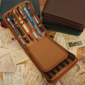 Puede incluir: Un estuche de cuero marrón con cremallera para bolígrafos y lápices. El estuche está abierto y tiene varios bolígrafos y lápices en su interior. El estuche está sobre una mesa con postales antiguas.