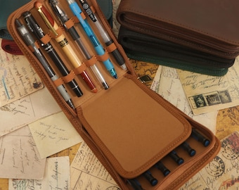 May include: A brown leather zippered case for pens and pencils. The case is open and has several pens and pencils inside. The case is sitting on a table with old postcards.