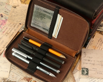 May include: A brown leather zippered pouch with a mesh pocket and elastic bands to hold pens and pencils. The pouch is open and has several pens and pencils inside.