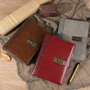 Op de afbeelding: Drie lederen dagboeken met cijfersloten. De dagboeken zijn bruin, rood en grijs. De dagboeken hebben decoratieve patronen op de omslagen.