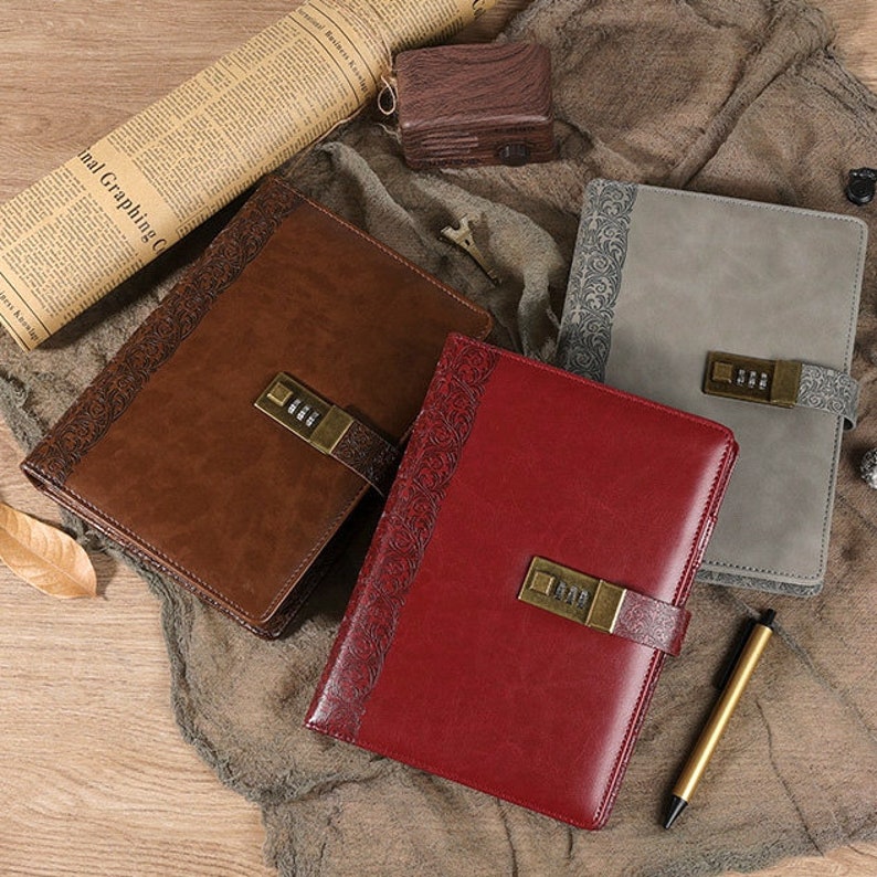 Op de afbeelding: Drie lederen dagboeken met cijfersloten. De dagboeken zijn bruin, rood en grijs. De dagboeken hebben decoratieve patronen op de omslagen.