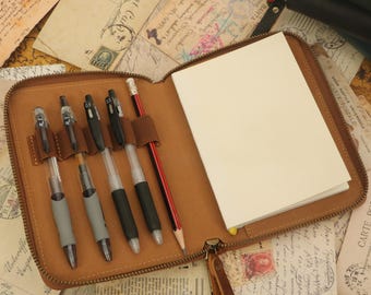 May include: A brown leather zippered notebook with a pen holder and a blank notebook inside. The notebook has a zipper closure and is made of brown leather. The pen holder has six slots for pens and pencils.