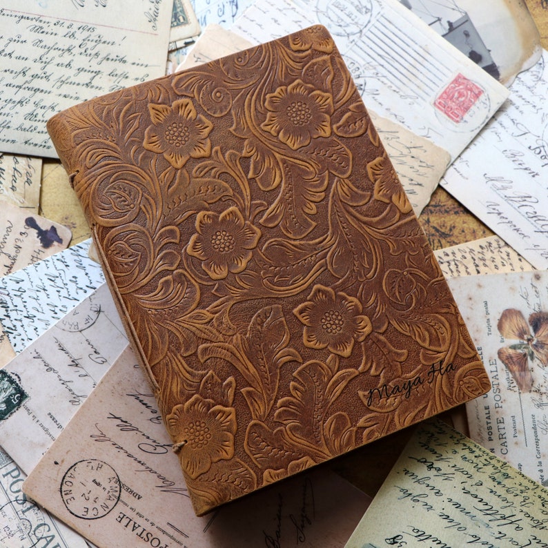 K&ouml;nnte beinhalten: Ein braunes Lederjournal mit einem gepr&auml;gten Blumenmuster. Das Journal tr&auml;gt den Namen "Maya Ha" auf der Vorderseite.
