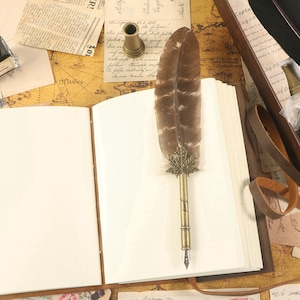 Puede incluir: Un diario encuadernado en cuero marr&oacute;n con p&aacute;ginas en blanco, una pluma con una pluma y una punta de metal, y un tintero de lat&oacute;n. La pluma tiene el nombre "Jenny" grabado en la punta de metal.