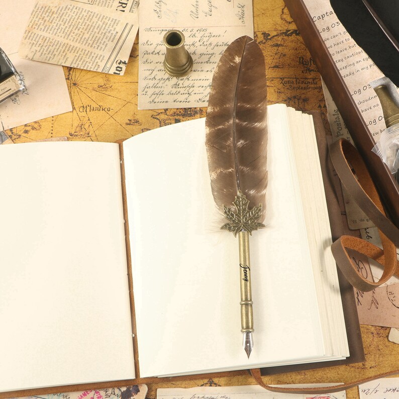 Puede incluir: Un diario encuadernado en cuero marr&oacute;n con p&aacute;ginas en blanco, una pluma con una pluma y una punta de metal, y un tintero de lat&oacute;n. La pluma tiene el nombre "Jenny" grabado en la punta de metal.