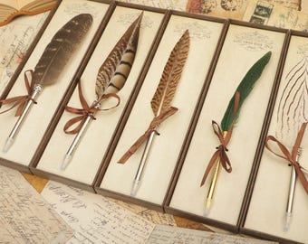 May include: Five feather pens in individual boxes with brown ribbon bows. The pens have different colored feathers, including brown, black, and green.