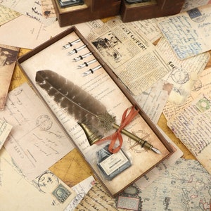 Puede incluir: Una pluma estilogr&aacute;fica de pluma marr&oacute;n y negra con una punta de metal y una punta de metal decorativa. La pluma est&aacute; en una caja con una peque&ntilde;a botella de tinta y varias puntas de plata. La caja est&aacute; rodeada de papel de aspecto vintage.