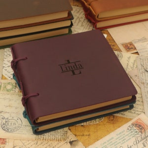 Może przedstawiać: Burgundowy skórzany dziennik z spersonalizowanym napisem "Linda" ze stylizowanym monogramem "L". Dziennik ma ozdobne zapięcie z paskiem i klamrą.