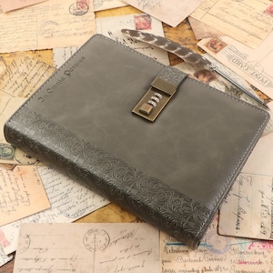 Op de afbeelding: Een grijs lederen gebonden dagboek met een combinatie slot en de inscriptie "In Omnia Paratus" op de voorzijde. Het dagboek is omringd door vintage ansichtkaarten.
