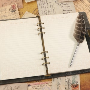 Op de afbeelding: Een zwart lederen gebonden dagboek met gouden ringen en een pen met een veer. Het dagboek heeft gelinieerde pagina's en de tekst "THE CLASSIC" is op de omslag gedrukt.