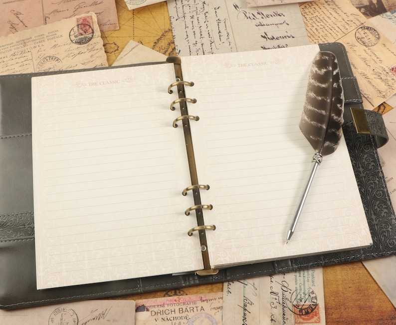 Op de afbeelding: Een zwart lederen gebonden dagboek met gouden ringen en een pen met een veer. Het dagboek heeft gelinieerde pagina's en de tekst "THE CLASSIC" is op de omslag gedrukt.