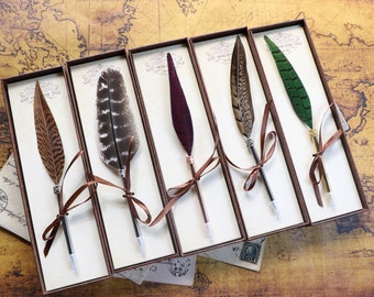 May include: Five feather pens in individual boxes with brown ribbon tied around each pen. The boxes are lined with white paper and have a label with the text "Vintage Quill Pen". The pens have different colored feathers, including brown, purple, green, and black.