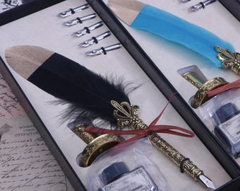 Pode incluir: Um conjunto de duas canetas de caligrafia com penas pretas e azuis, frascos de tinta e pontas em uma caixa preta. As canetas têm detalhes ornamentados em metal dourado. Os frascos de tinta são etiquetados como "The Feathered Pen".