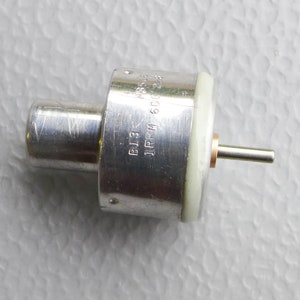 May include: A silver electric motor with a white plastic gear. The motor has a shaft with a small metal pin. The motor has the text "1 RPM 6000", "B13", and "JRPN" printed on the side.